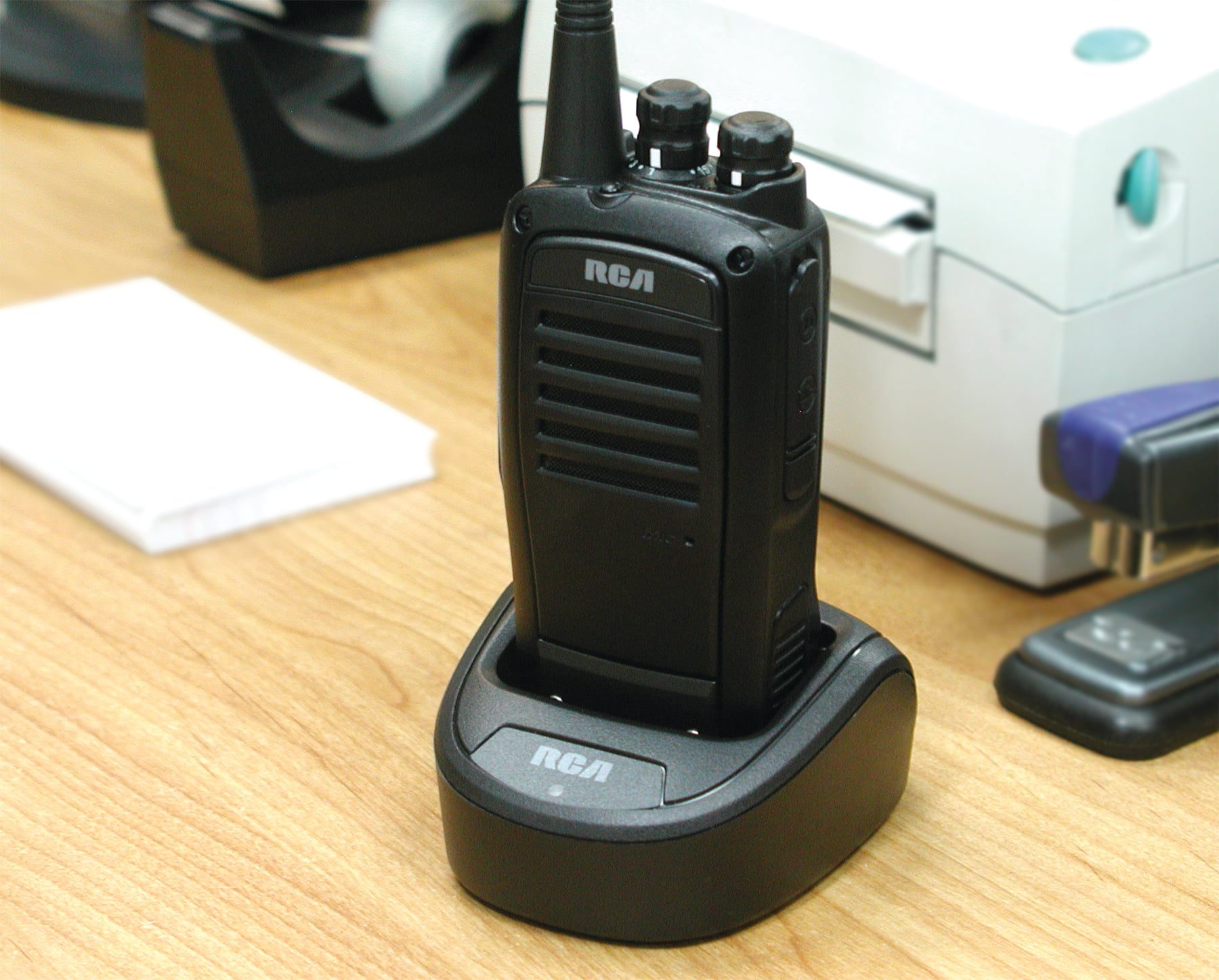 An RCA 2-way handheld radio rests in its charger on a desk.