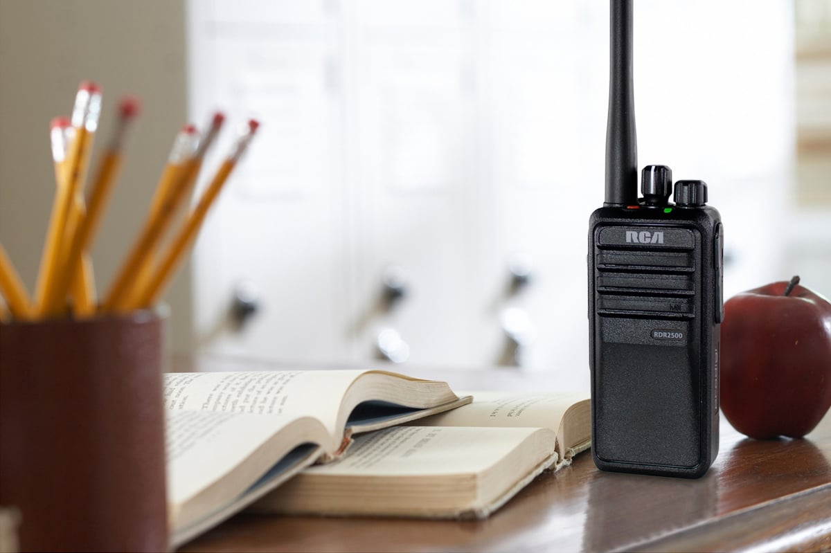 An RCA RDR2500 school two-way radio is set upright on a teacher's desk beside two textbooks, an apple, and a pencil holder.